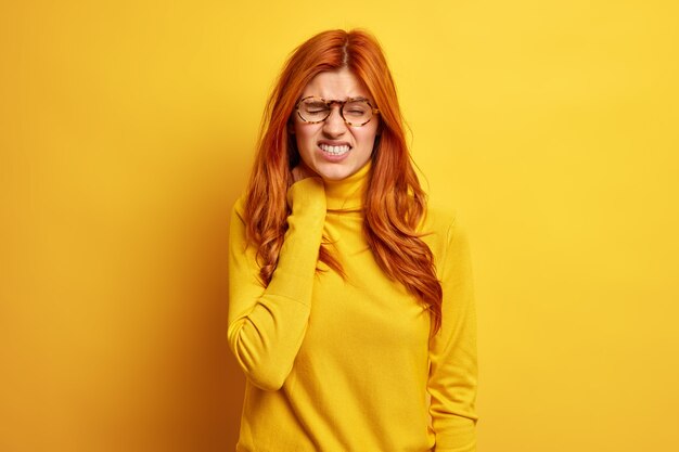 Redhead displeased woman touches neck feels strong pain has office syndrom leads sedentary lifestyle wears spectacles turtleneck.
