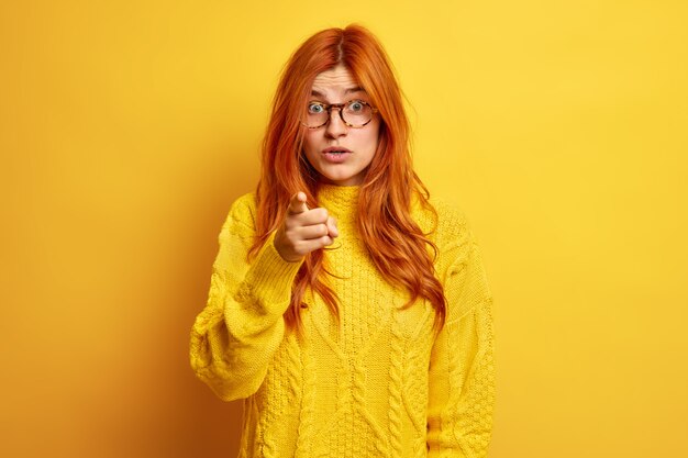 Redhead European woman indicates at you with index finger feels shocked to see something amazing wears transparent glasses and warm sweater.