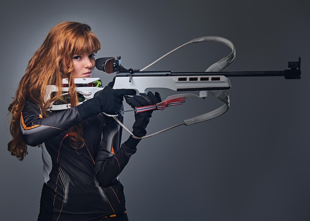 Redhead female Biathlon champion aiming with a competitive gun in a studio over grey background.Redhead female Biathlon champion aiming with a competitive gun in a studio over grey background.