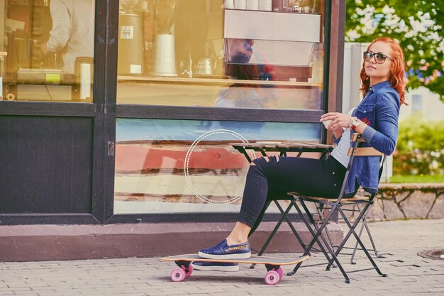 빨간 머리 여성은 Longboard에서 스케이트를 탄 후 커피를 마십니다.