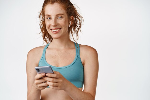 Рыжая фитнес-девушка с помощью приложения для смартфона слушает музыку в беспроводных наушниках и улыбается на тренировке с музыкой, стоящей на белом фоне