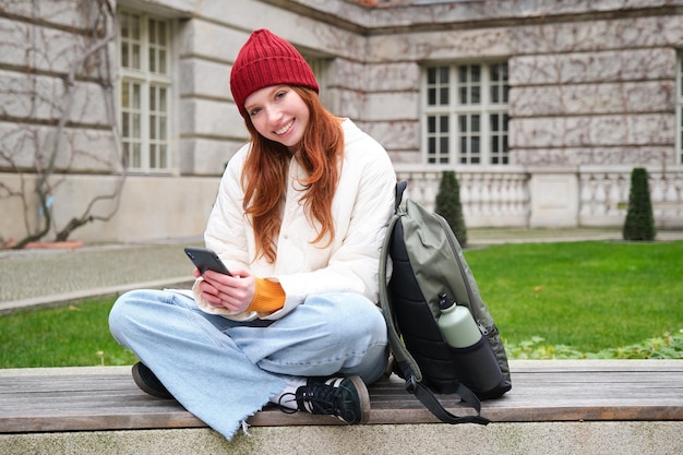 Бесплатное фото Рыжая студентка сидит с мобильным телефоном на скамейке в парке, опираясь на свой рюкзак