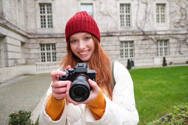 Foto gratuita il fotografo di ragazze rosse scatta foto con una fotocamera professionale all'aperto cattura scatti streetstyle lo