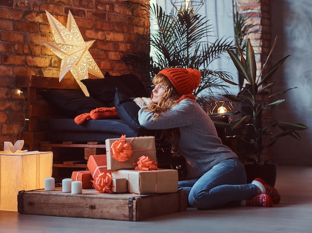 床に座っているセーターと帽子をかぶった赤毛の女の子は、クリスマスの時期に装飾されたリビングルームの窓の外を夢のように見ています。