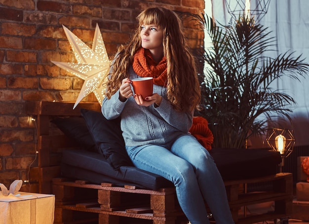 Redhead girl wearing a sweater sitting on a couch, warming with a cup of coffee in a decorated living room at Christmas time.