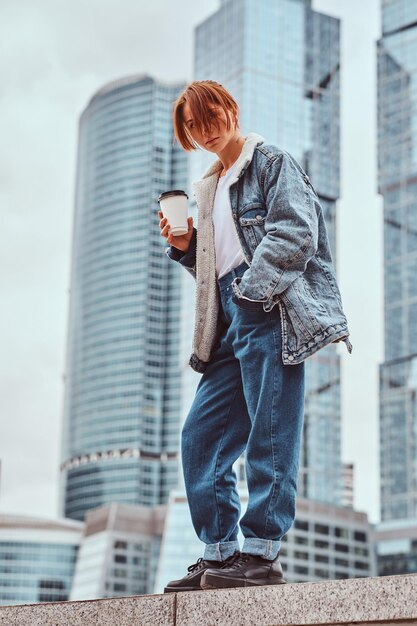 모스크바 시의 고층 빌딩 앞에서 테이크아웃 커피를 들고 데님 코트를 입고 얼굴에 문신을 한 빨간 머리 힙스터 소녀.