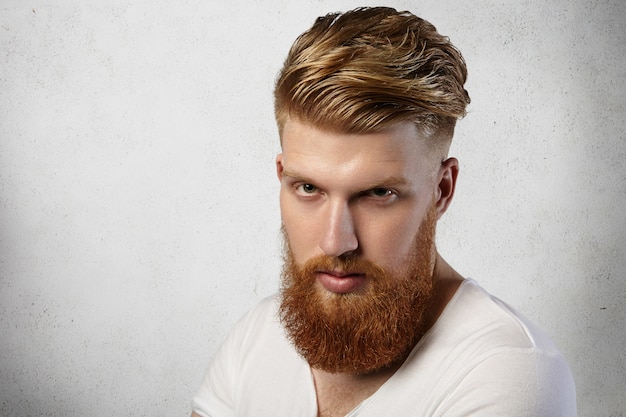 Redhead hipster with trendy haircut and fuzzy beard dressed in white t-shirt posing indoors, having serious and sullen look.