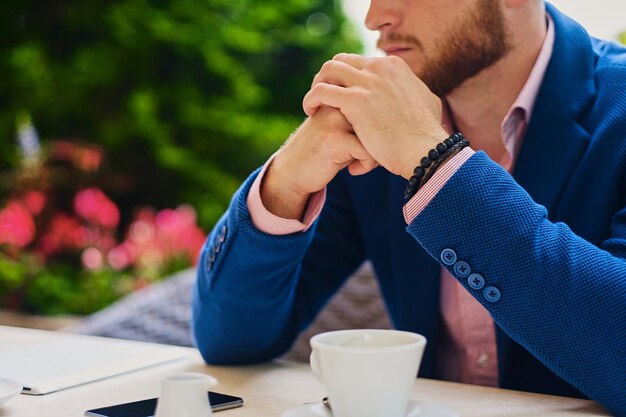 A redhead male dressed in a pink shirt, drinks coffee in an open air cafe.