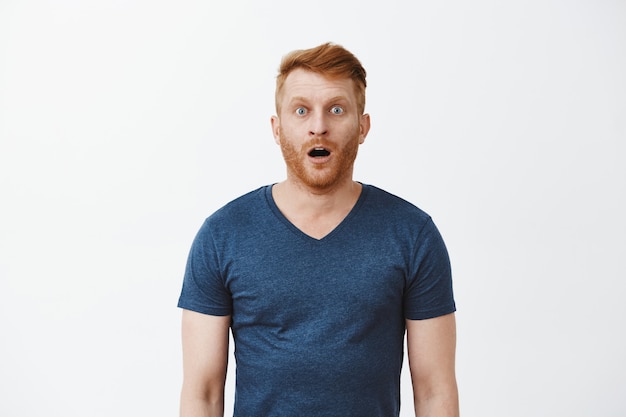 Redhead male with bristle dropping jaw as seeing incredible and amazing scenery, posing over gray wall in casual blue t-shrit, gasping, staring thrilled and astonished over grey wall