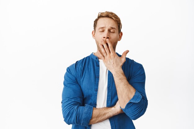 Redhead man feels tired and yawns, covers mouth with palm and close eyes, exhausted or bored, stands against white wall in casual clothes