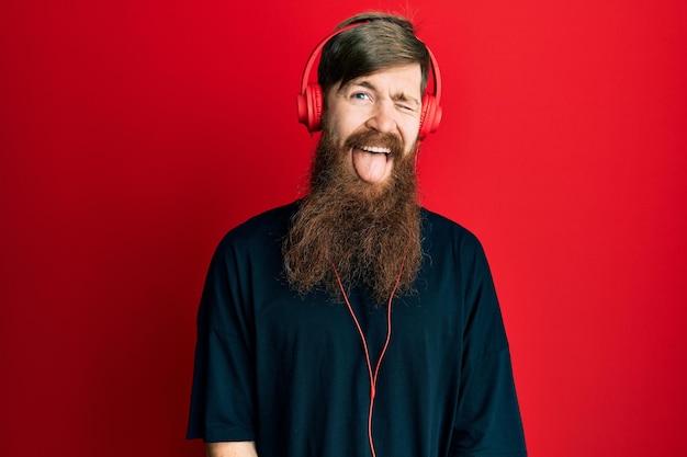 Рыжий мужчина с длинной бородой слушает музыку в наушниках, подмигивая, глядя в камеру с сексуальным выражением лица, веселым и счастливым лицом.