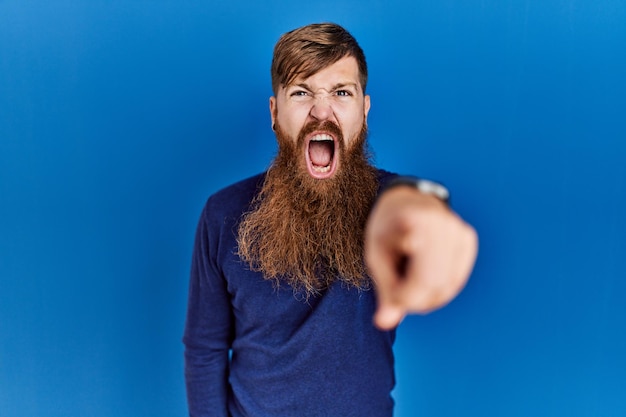 Красноволосый мужчина с длинной бородой в обычном синем свитере на синем фоне, указывающий недовольно и разочарованно на камеру, злится и в ярости на тебя.