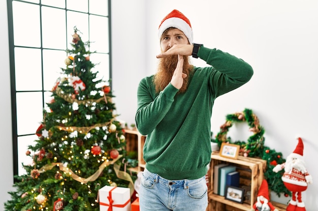 Uomo rosso con la barba lunga che indossa il cappello di natale vicino all'albero di natale che fa un gesto di timeout con le mani, viso frustrato e serio