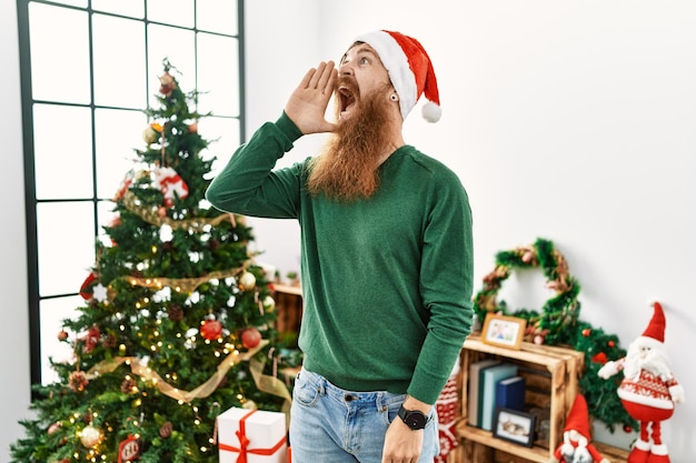 Redhead man with long beard wearing christmas hat by christmas tree shouting and screaming loud to side with hand on mouth communication concept