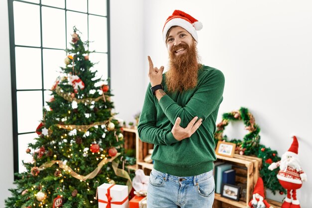 긴 수염을 가진 빨간 머리 남자는 크리스마스 트리 옆에서 크리스마스 모자를 쓰고 카메라를 바라보며 손 손가락으로 옆을 가리키는 얼굴에 큰 미소를 지었다