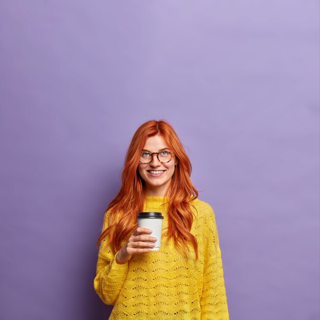 redhead millennial woman holds takeaway coffee smiles positively dressed in yellow jumper.