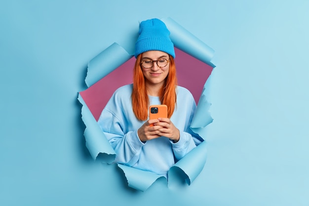 Рыжая довольная молодая кавказская женщина использует мобильный телефон, набирает смс-сообщения в социальных сетях, носит синюю шляпу и толстовку.