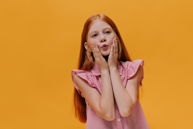 Redhead pretty girl in red striped shirt touches her cheeks and whistles Charming child poses on orange isolated background