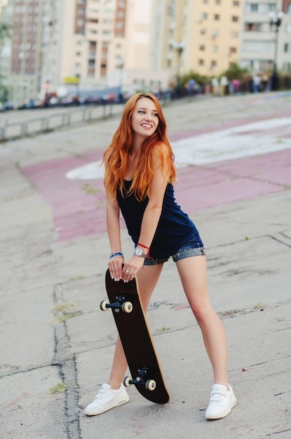 ジーンズのショートパンツと都市の科学でスケートボードでポーズをとる黒のtシャツの赤毛のスリムな女の子。