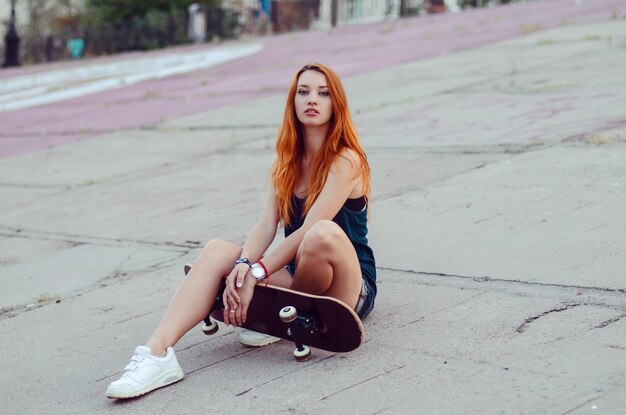 Redhead slim girl in jeans shorts and black t shirt posing with skateboard in urban sciene.