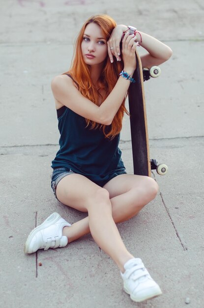 Redhead slim girl in jeans shorts and black t shirt posing with skateboard in urban sciene.