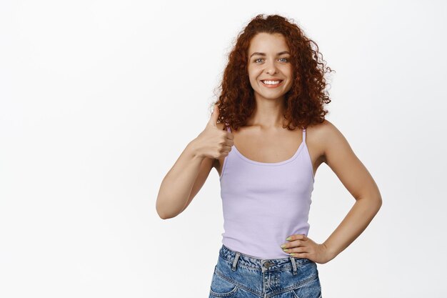 Redhead smiling woman shows thumbs up, looking happy and motivated, complimenting, standing in tank top against white background.