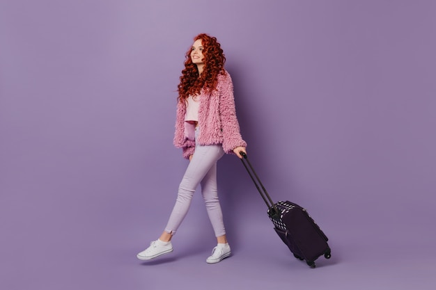Redhead traveler girl in pink coat and white jeans comes with suitcase on purple space.
