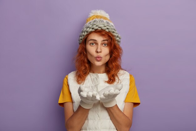 Redhead woman blows at camera, keeps palms forward, wears knitted hat and white vest, enjoys winter time