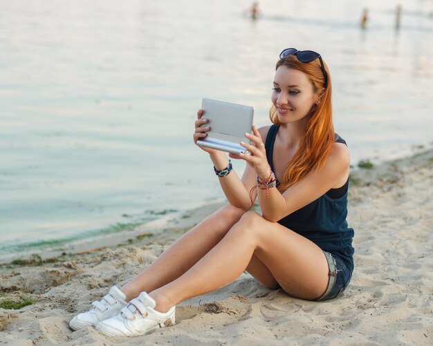 Redhead woman in jeans shorts and blue t shirt sitting on a beach and holding tablet computer.