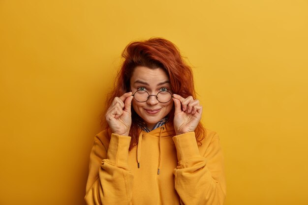 Redhead woman looks scrupulously through optical glasses, has curious gaze, keeps hands on frame of spectacles, wears yellow sweatshirt