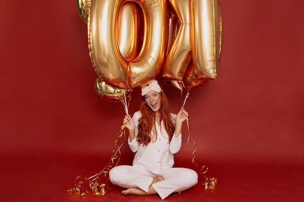 redhead woman in pijama and sleeping mask posing excited holding golden balloons on red