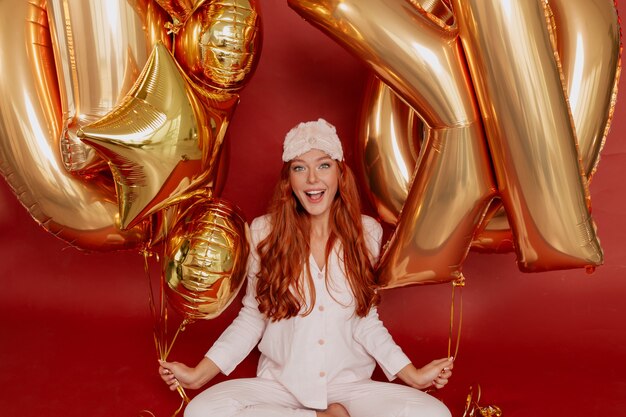 redhead woman in pijama and sleeping mask posing excited holding golden balloons on red