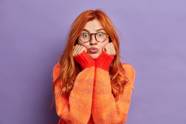 Foto gratuita donna rossa con espressione sorpresa tiene le mani sotto il mento tiene le labbra piegate vestita con un maglione casual fa una smorfia divertente.