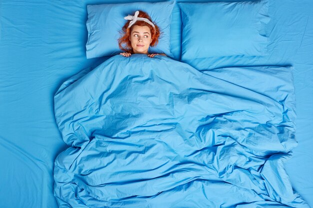 redhead young European woman lying under soft blanket wears headband sleeps in cozy bedroom enjoys good morning feels comfortable wears headband looks thoughtfully aside plans day