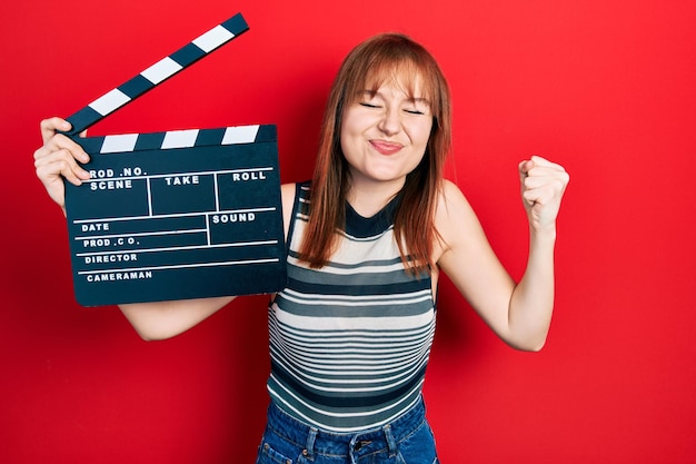 Redhead young woman holding video film clapboard screaming proud, celebrating victory and success very excited with raised arm