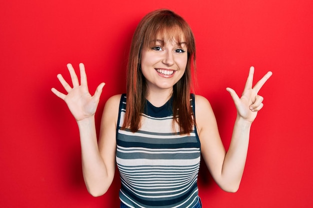 無料写真 カジュアルなtシャツを着た赤毛の若い女性が、自信と幸せな笑みを浮かべながら、指8番を見せて上向きにしている。
