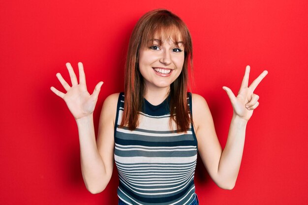 カジュアルなTシャツを着た赤毛の若い女性が、自信と幸せな笑みを浮かべながら、指8番を見せて上向きにしている。