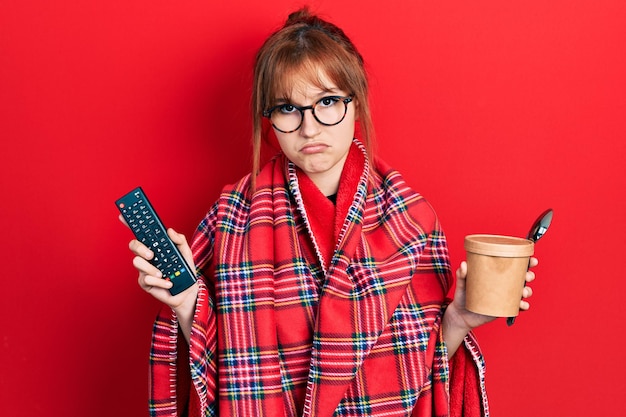 Redhead young woman wrapped in a red warm red blanket eating icecream and watching tv depressed and worry for distress crying angry and afraid sad expression