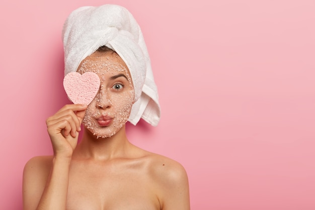 Reducing pores and cleansing concept. Attractive female applies sea salt mask on face, has luxurious feelings from beauty treatments, covers eye with heart shaped sponge, pampers complexion.