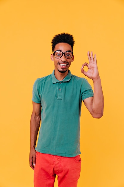 Refined african boy in red pants posing with okay sign. Positive guy in big trendy glasses standing.