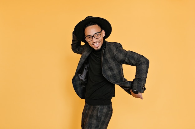 Refined african man having fun during photoshoot . Indoor photo of smiling tall black guy in hat and glasses dancing on yellow wall.