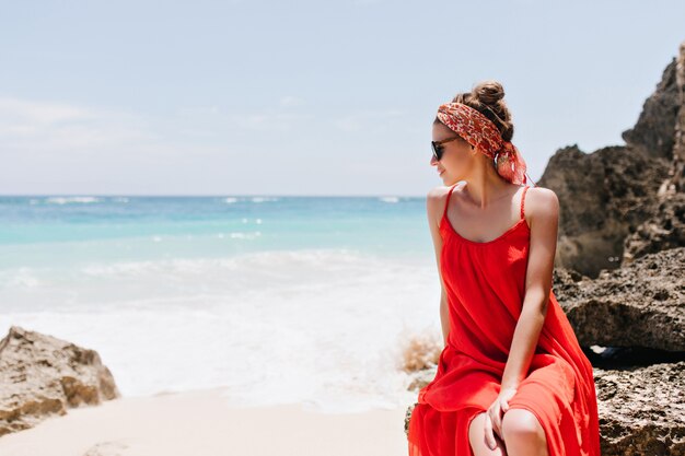 세련된 백인 여성 모델이 돌에 앉아 바다 전망을 즐기고 있습니다. 선글라스를 통해 바다를보고 로맨틱 백인 젊은 여자.