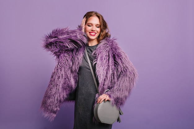 Refined curly girl with trendy make-up standing with eyes closed on purple background