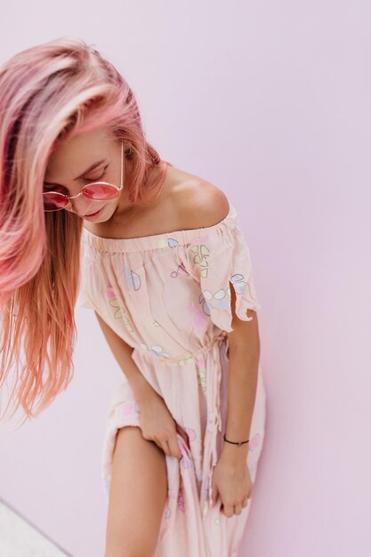 Refined european model with tanned skin looking down during photoshoot in long dress Blissful pinkhaired girl in trendy sunglasses standing beside white wall