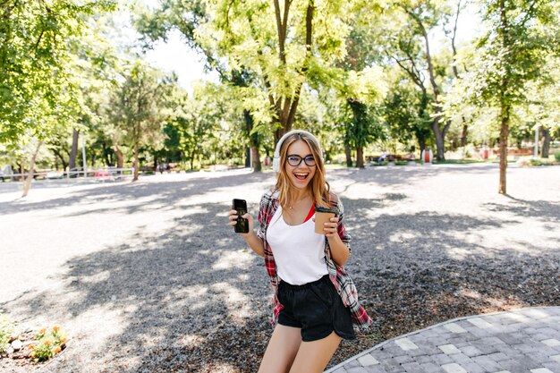 公園で笑っている黒のショートパンツで洗練された金髪の女性。夏の週末に一杯のコーヒーが浮かんでいる壮大なヨーロッパの女の子。