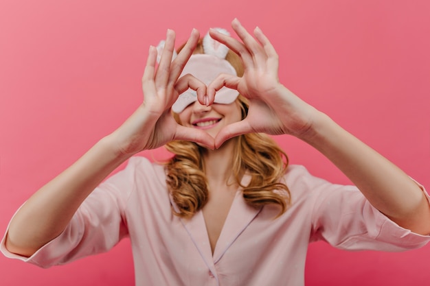 Foto gratuita raffinata donna bionda in maschera da skeep scherzare al mattino. allegra ragazza caucasica in tuta da notte che fa il segno del cuore.