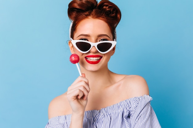 Refined ginger woman holding hard candy and laughing. Studio shot of glad pinup girl in sunglasses isolated on blue space.