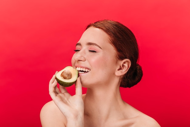 Refined girl eating avocado Studio shot of enchanting caucasian woman posing on red background