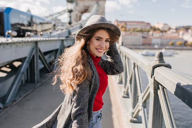 Refined girl in elegant tweed coat posing with charming smile on urban background during travel