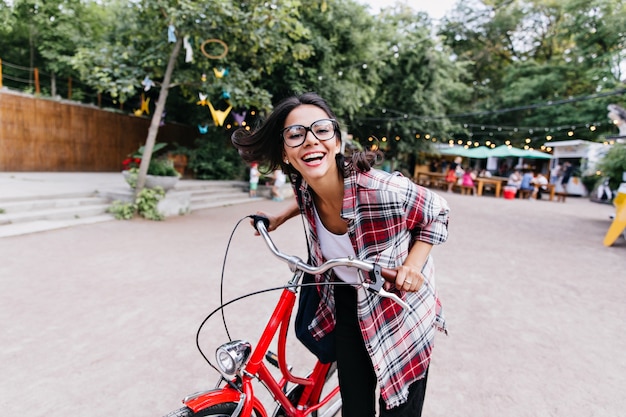 Refined girl in stylish glasses riding around town. Outdoor photo of lovely dark-haired woman sitting on bicycle in front of trees.